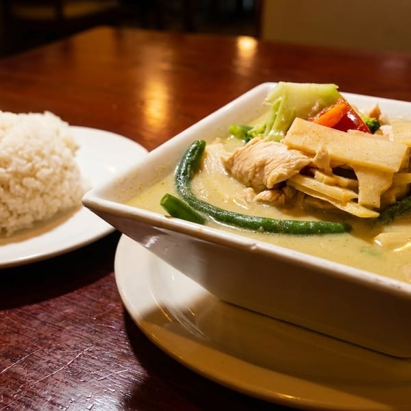 Green curry with chicken, bamboo shoots, and vegetables, served with white rice on a wooden table.